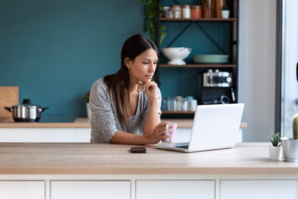 mujer joven mirando su portátil mientras sostiene una taza de café en la cocina